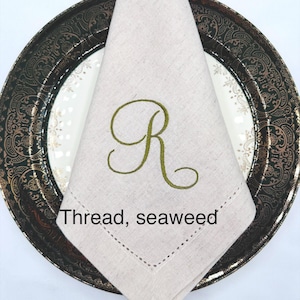 May include: A folded beige linen napkin with a green embroidered letter "R" sits on a decorative plate. The plate has a dark rim with gold accents. The text "Thread, seaweed" is visible.