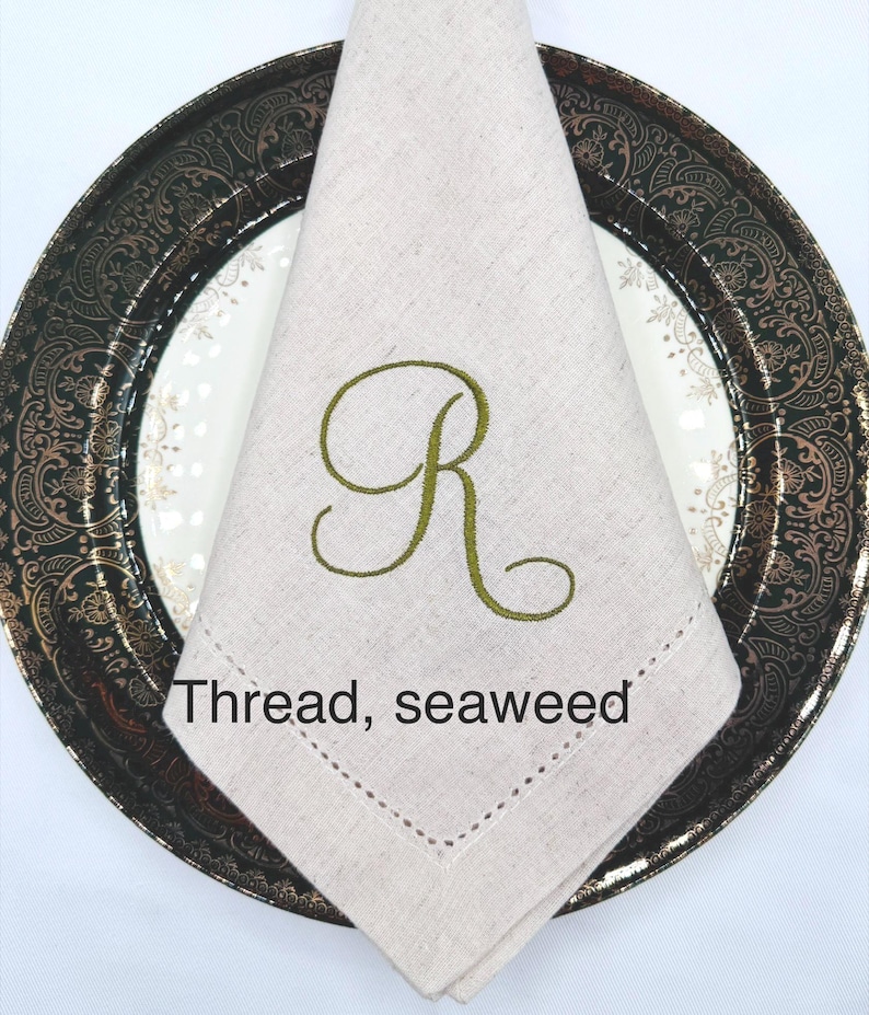 May include: A folded beige linen napkin with a green embroidered letter "R" sits on a decorative plate. The plate has a dark rim with gold accents. The text "Thread, seaweed" is visible.