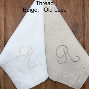May include: Two folded linen napkins, one cream and one beige, each embroidered with the letter "R" in a matching thread color. The napkins have a stitched border and are displayed on a wooden surface.