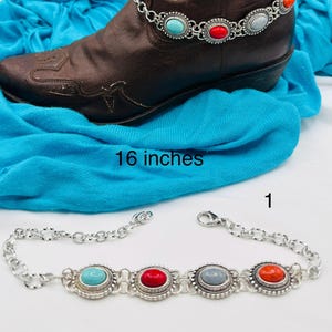 May include: A silver-toned chain boot bracelet featuring four oval cabochon stones in turquoise, red, grey, and orange, each set in a decorative metal frame. The bracelet measures 16 inches in length and is shown wrapped around a brown leather boot.