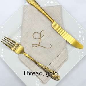 May include: A gold-colored fork and knife with ornate floral detailing, placed on a white square plate. A beige linen napkin with a gold embroidered "L" is beneath the cutlery. The text "Thread, gold" is visible.