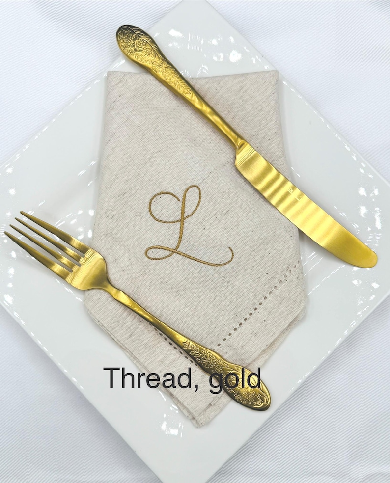 May include: A gold-colored fork and knife with ornate floral detailing, placed on a white square plate. A beige linen napkin with a gold embroidered "L" is beneath the cutlery. The text "Thread, gold" is visible.