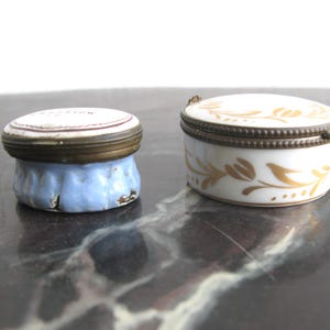 May include: Two small antique porcelain trinket boxes. One is a round, white box with gold floral designs and a bronze-coloured metal rim. The other is a small, round box with a blue base and a white lid with text.
