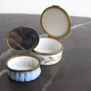 Puede incluir: Dos peque&ntilde;as cajas de baratijas antiguas. Una est&aacute; abierta, revelando un espejo y un interior azul. La otra es blanca con detalles florales dorados y una tapa con bisagras. Ambas tienen bordes de metal dorado.