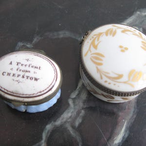 Puede incluir: Dos joyeros antiguos de porcelana. Uno es redondo con tapa blanca y dise&ntilde;o floral dorado. El otro es ovalado con el texto "A Present from CHEPSTOW" en la tapa. Ambos tienen bordes met&aacute;licos.