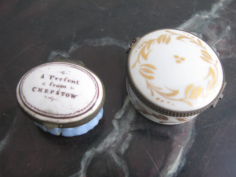 Puede incluir: Dos joyeros antiguos de porcelana. Uno es redondo con tapa blanca y dise&ntilde;o floral dorado. El otro es ovalado con el texto "A Present from CHEPSTOW" en la tapa. Ambos tienen bordes met&aacute;licos.