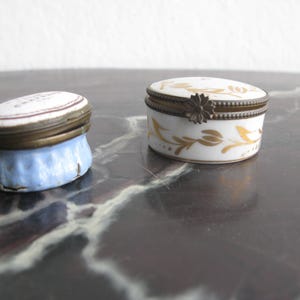 Puede incluir: Dos peque&ntilde;as cajas de baratijas antiguas. Una tiene una base azul claro con tapa blanca y ribete de lat&oacute;n. La otra es blanca con dise&ntilde;os florales dorados y cierre de lat&oacute;n. Ambas son de forma ovalada.