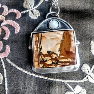 Può includere: Una collana con pendente in argento con una pietra rettangolare con motivi marroni e crema. Il pendente ha una piccola pietra bianca rotonda e pende da una catena d'argento. Lo sfondo è un tessuto scuro con ricami floreali.