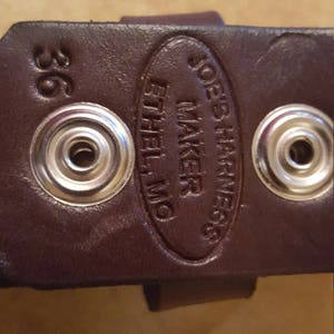May include: Close-up of a brown leather belt with a silver buckle. The belt is stamped with "Joe's Harness Maker Ethel, MO" and "36".
