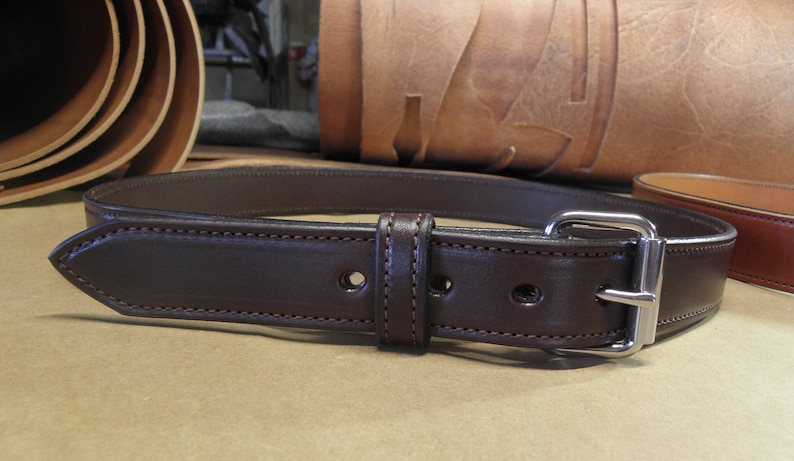 May include: A brown leather belt with a silver buckle. The belt is stitched with a brown thread and has two rows of stitching.