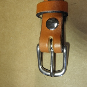 May include: Close-up of a brown leather belt with a silver buckle. The buckle has a smooth, rounded finish and a simple design.