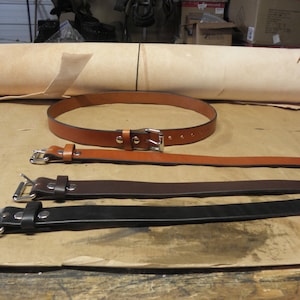 May include: Three leather belts with silver buckles. The belts are brown, dark brown, and black. A roll of light brown leather is in the background.
