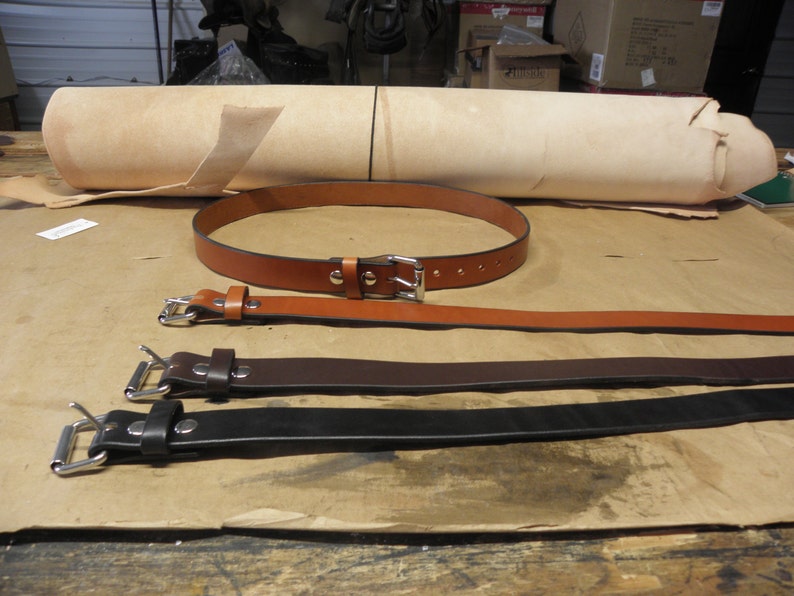 May include: Three leather belts with silver buckles. The belts are brown, dark brown, and black. A roll of light brown leather is in the background.