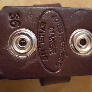 May include: Close-up of a brown leather belt with metal snaps and the text "Joe's Harness Maker Ethel, MO" embossed on the leather.