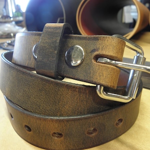 May include: A brown leather belt with a silver buckle. The belt is folded in half and the buckle is visible. The belt is made of a distressed leather.