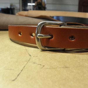 May include: A brown leather belt with a silver buckle. The belt has multiple holes for adjusting the fit.