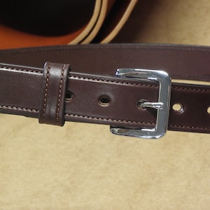 May include: A brown leather belt with a silver buckle. The belt has a stitched edge and multiple holes for adjusting the size.