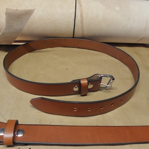 May include: A brown leather belt with a silver buckle and five belt loops. The belt is rolled up and laid on a brown surface.