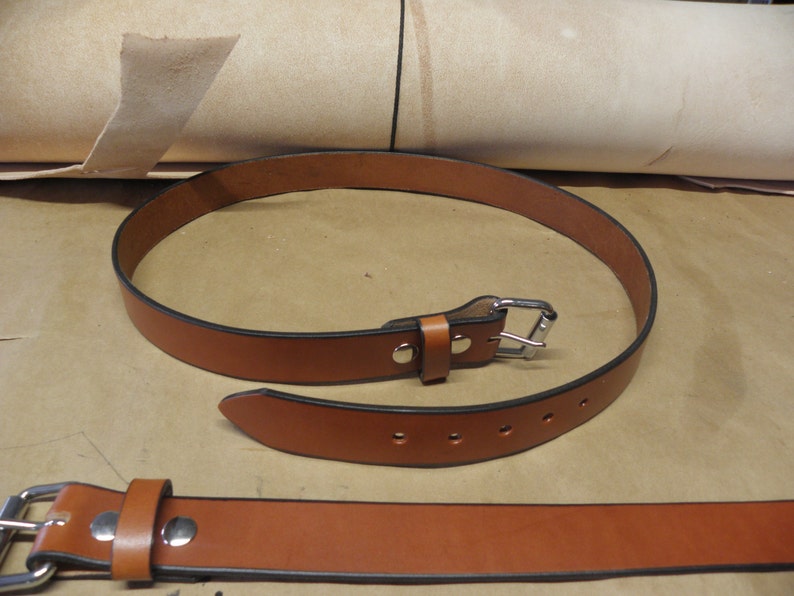 May include: A brown leather belt with a silver buckle and five belt loops. The belt is rolled up and laid on a brown surface.