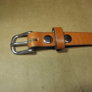 May include: A brown leather belt with a silver buckle and two silver rivets. The belt is folded in half and the buckle is closed.