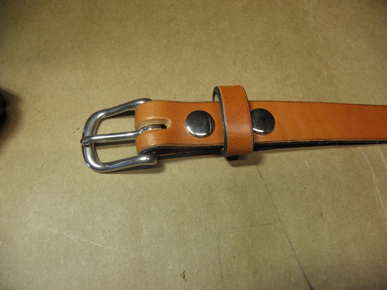 May include: A brown leather belt with a silver buckle and two silver rivets. The belt is folded in half and the buckle is closed.