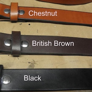 May include: Three leather belts in different colors: chestnut, British brown, and black. Each belt has a metal buckle and is shown lying flat on a light brown surface.