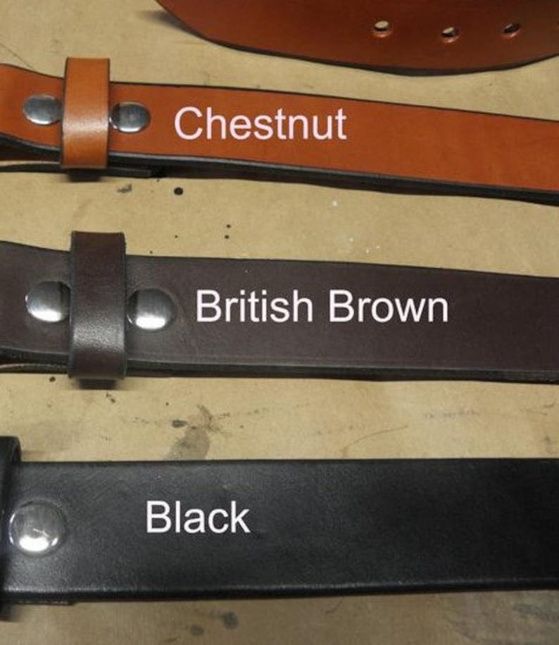 May include: Three leather belts in different colors: chestnut, British brown, and black. Each belt has a metal buckle and is shown lying flat on a light brown surface.