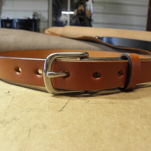 May include: A brown leather belt with a silver buckle. The belt has five holes for adjusting the size.
