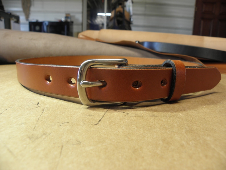 May include: A brown leather belt with a silver buckle. The belt has five holes for adjusting the size.