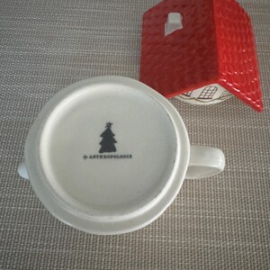 K&ouml;nnte beinhalten: Eine wei&szlig;e Keramiktasse mit Henkel, mit einem schwarzen Weihnachtsbaum-Design und dem Text "by ANTHROPOLOGIE" auf der Unterseite. Ein roter Keramikdeckel in Hausform befindet sich neben der Tasse.