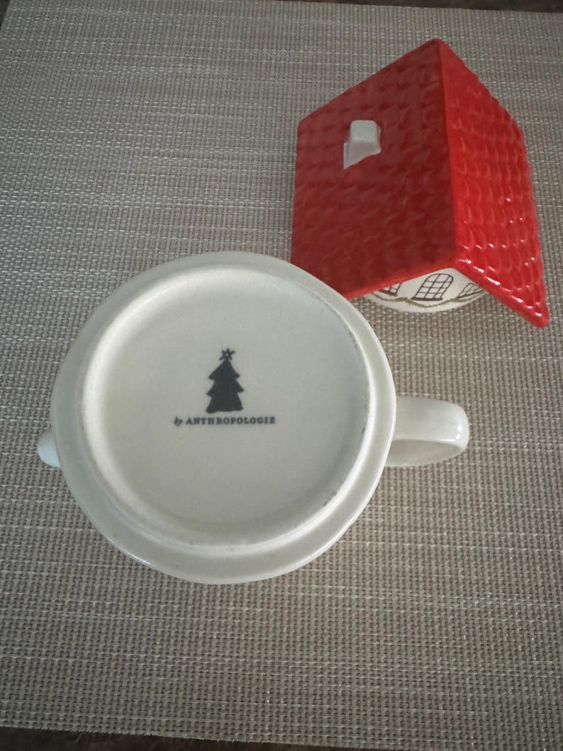 K&ouml;nnte beinhalten: Eine wei&szlig;e Keramiktasse mit Henkel, mit einem schwarzen Weihnachtsbaum-Design und dem Text "by ANTHROPOLOGIE" auf der Unterseite. Ein roter Keramikdeckel in Hausform befindet sich neben der Tasse.