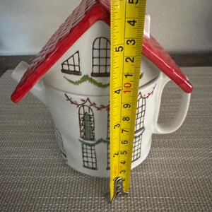 K&ouml;nnte beinhalten: Eine wei&szlig;e Keramik-Teekanne in Form eines Hauses mit rotem Dach. Die Teekanne hat schwarze Fensterdetails und gr&uuml;ne Akzente. Die H&ouml;he der Teekanne betr&auml;gt ca. 12.7 cm.