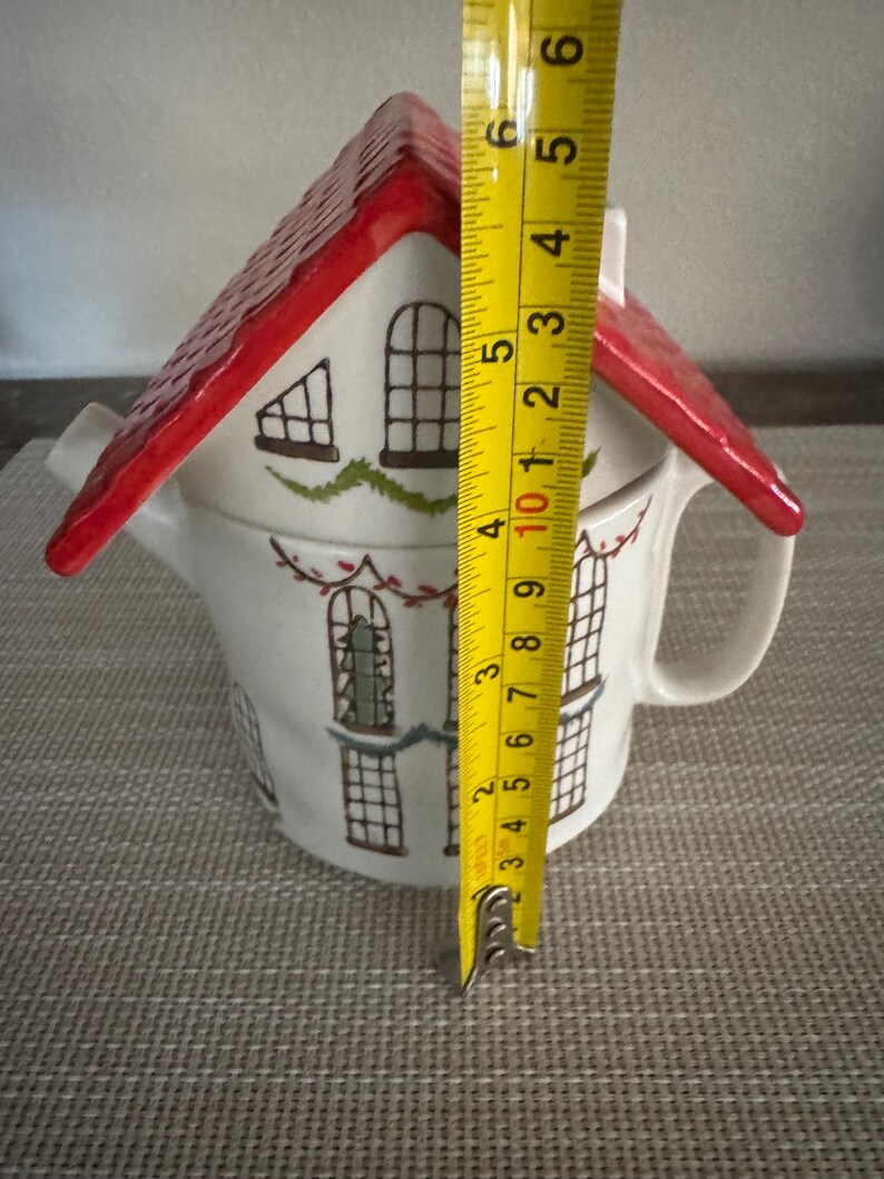 K&ouml;nnte beinhalten: Eine wei&szlig;e Keramik-Teekanne in Form eines Hauses mit rotem Dach. Die Teekanne hat schwarze Fensterdetails und gr&uuml;ne Akzente. Die H&ouml;he der Teekanne betr&auml;gt ca. 12.7 cm.