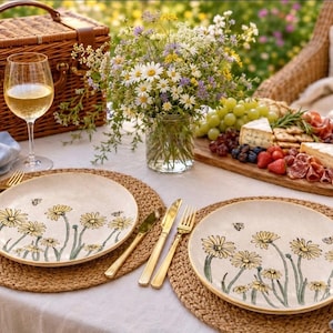 May include: Two round ceramic plates featuring hand-painted yellow daisy flowers and small bees on a cream background. The plates are set on woven jute placemats with gold-toned cutlery, light blue linen napkins, and a picnic basket in the background.