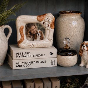 May include: A decorative shelf arrangement featuring a dog-themed picture frame, a ceramic dog figurine, and stacked books. The frame holds a photo of a woman and a dog. The books read "PETS ARE MY FAVORITE PEOPLE" and "ALL YOU NEED IS LOVE AND A DOG."