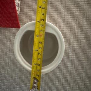 K&ouml;nnte beinhalten: Ein wei&szlig;er Keramikbeh&auml;lter mit ovaler &Ouml;ffnung, ca. 10 cm breit. Ein gelbes Ma&szlig;band zeigt die Abmessungen. Ein rotes Keramikhaus ist im Hintergrund zu sehen.