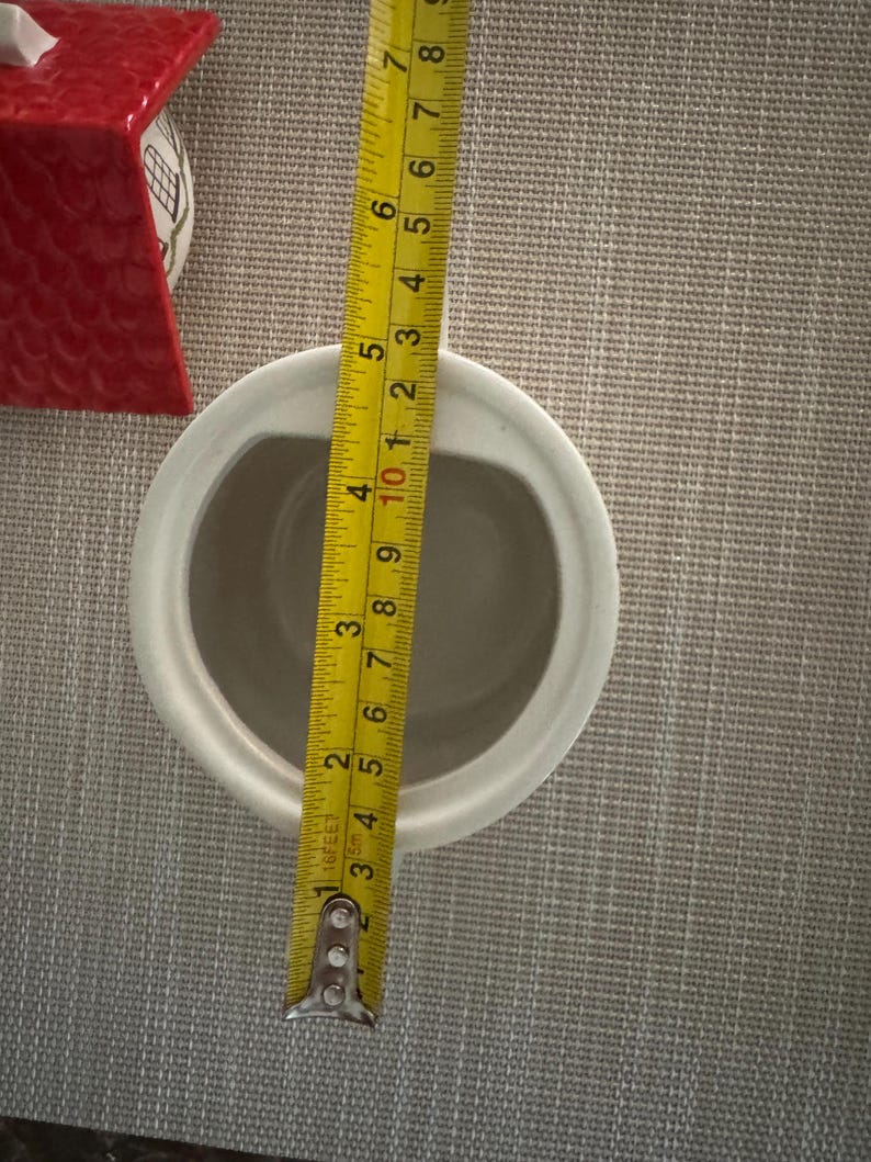 K&ouml;nnte beinhalten: Ein wei&szlig;er Keramikbeh&auml;lter mit ovaler &Ouml;ffnung, ca. 10 cm breit. Ein gelbes Ma&szlig;band zeigt die Abmessungen. Ein rotes Keramikhaus ist im Hintergrund zu sehen.