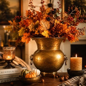 May include: A large, hammered brass urn with lion head ring handles, filled with an arrangement of orange and yellow autumn leaves and berries. Beside it are a glass candle holder, a bowl of small pumpkins, and a pillar candle on a dark base.