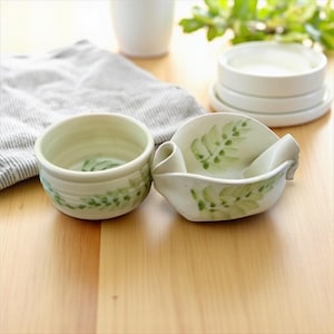 May include: Two ceramic bowls with a hand-painted green leaf design. One is a round bowl, and the other has a unique shape with a handle. The bowls are off-white and sit on a wooden surface.