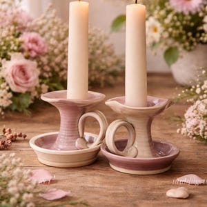 May include: Two lit ivory candles in decorative ceramic candle holders with handles. The candle holders are two-toned, with a light purple and cream color scheme. The background features pink flowers and greenery.