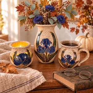 May include: A ceramic set featuring a speckled beige vase, a candle holder, and a mug, each decorated with hand-painted blue iris flowers and green leaves. The vase holds a bouquet of autumn-colored leaves, berries, and blue flowers.