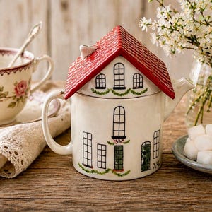 K&ouml;nnte beinhalten: Eine wei&szlig;e Keramik-Teekanne in Form eines Hauses mit rotem Dach und schwarzen Fensterdetails. Eine blumige Teetasse, Untertasse, eine Spitzenserviette und ein Teller mit Zuckerw&uuml;rfeln sind ebenfalls zu sehen.