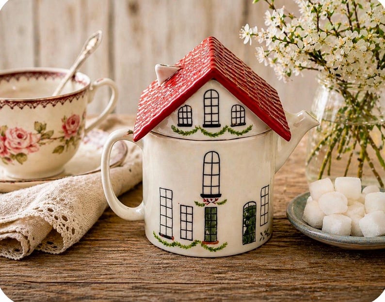K&ouml;nnte beinhalten: Eine wei&szlig;e Keramik-Teekanne in Form eines Hauses mit rotem Dach und schwarzen Fensterdetails. Eine blumige Teetasse, Untertasse, eine Spitzenserviette und ein Teller mit Zuckerw&uuml;rfeln sind ebenfalls zu sehen.