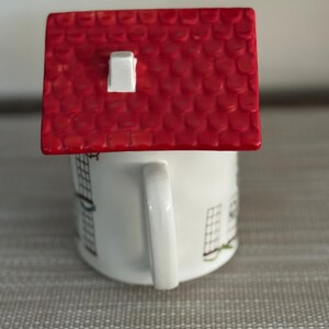 K&ouml;nnte beinhalten: Eine wei&szlig;e Keramiktasse mit einem roten dachf&ouml;rmigen Deckel. Die Tasse hat einen Henkel und ist mit einem Hausdesign verziert. Das Dach ist leuchtend rot mit einem wei&szlig;en Schornstein.