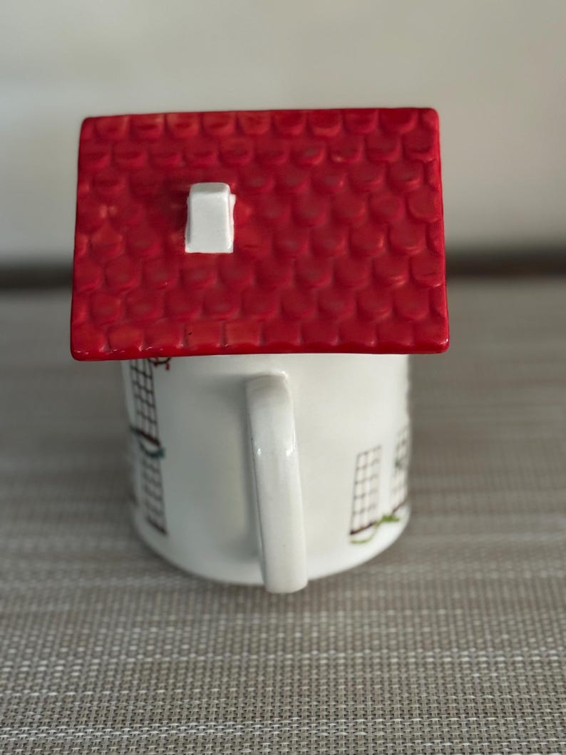 K&ouml;nnte beinhalten: Eine wei&szlig;e Keramiktasse mit einem roten dachf&ouml;rmigen Deckel. Die Tasse hat einen Henkel und ist mit einem Hausdesign verziert. Das Dach ist leuchtend rot mit einem wei&szlig;en Schornstein.