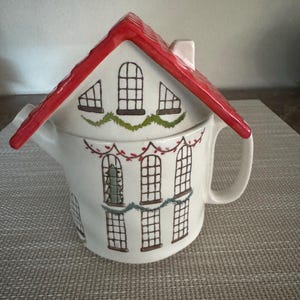 K&ouml;nnte beinhalten: Wei&szlig;e Keramik-Teekanne in Form eines Hauses mit rotem Dach. Die Teekanne hat schwarze Fensterdesigns und gr&uuml;ne Girlandenakzente. Ein kleiner Schornstein ist auf dem Dach sichtbar. Der Griff ist wei&szlig; und gebogen.