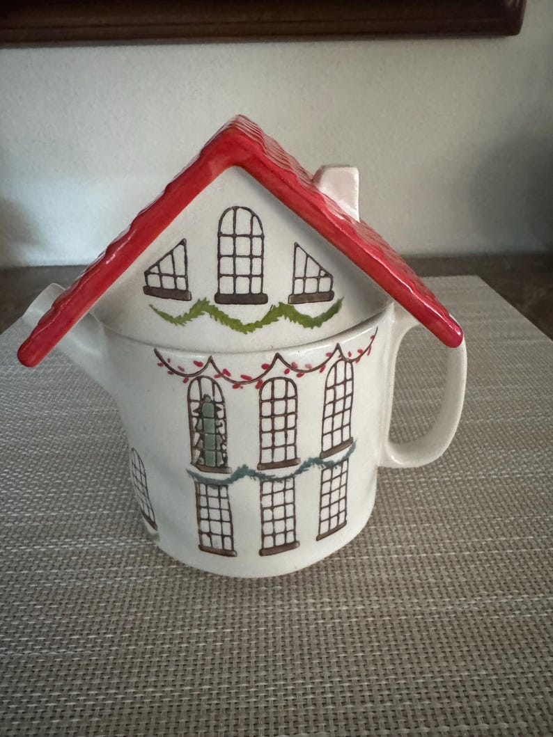 K&ouml;nnte beinhalten: Wei&szlig;e Keramik-Teekanne in Form eines Hauses mit rotem Dach. Die Teekanne hat schwarze Fensterdesigns und gr&uuml;ne Girlandenakzente. Ein kleiner Schornstein ist auf dem Dach sichtbar. Der Griff ist wei&szlig; und gebogen.