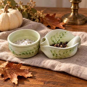 May include: A pair of light green ceramic bowls featuring hand-painted green leaf motifs. One bowl is round and filled with white salt crystals, while the other is an irregular, organic shape containing dark whole peppercorns.