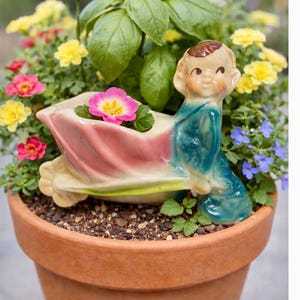May include: A terracotta flower pot filled with colorful flowers and greenery. A ceramic planter in the shape of a seated child is nestled among the plants. A ladybug decoration sits atop a stem.