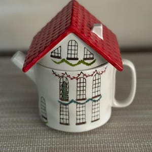 K&ouml;nnte beinhalten: Eine wei&szlig;e Keramik-Teekanne in Form eines Hauses mit rotem Dach. Die Teekanne hat bemalte Fenster und dekorative Girlanden. Henkel und Ausguss sind sichtbar, und das Design hat ein gem&uuml;tliches, festliches Thema.